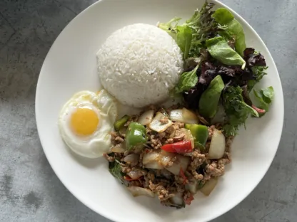 Thai basil stir-fry with ground chicken, onions, and bell peppers on a plate with jasmine rice, fried egg, and mixed greens.