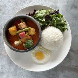 Thai red curry with tofu and vegetables in a bowl, alongside jasmine rice, fried egg, and greens arranged on a plate.