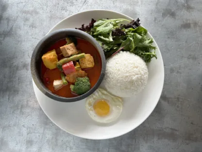 Thai red curry with tofu and vegetables in a bowl, alongside jasmine rice, fried egg, and greens arranged on a plate.