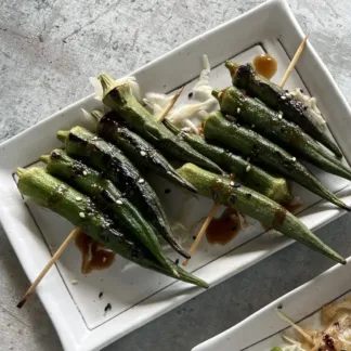 Grilled okra on wood skewers, topped with sesame seeds and teriyaki sauce on white plate