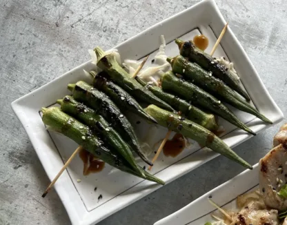 Grilled okra on wood skewers, topped with sesame seeds and teriyaki sauce on white plate