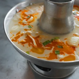 Coconut chicken soup with Enoki mushrooms, green onions, yellow onions, topped with chili oil, served in a metal hot pot.