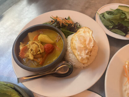 Thai yellow curry with chicken and vegetables served in a bowl. Rice topped with a fried egg and a side greens on a plate.